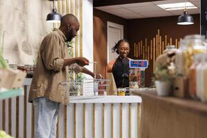 African American male customer unloading his shopping basket at the cashier counter. The friendly female vendor waits patiently with a smile and assists with the weighing of the products. photo