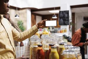 At eco friendly grocery shop African American male consumer holds a cell phone that displays an isolated white screen. Image shows black man with mobile device having a chromakey template. photo