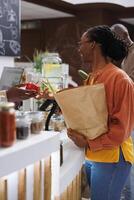 imagen de negro mujer con un tienda de comestibles bolso entrega terminado rojo pimienta a el africano americano local proveedor. un sonriente hembra cliente dando su seleccionado Fresco Produce a el cajero, sostenible compras. foto