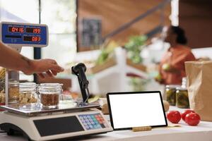 Glass jar filled with bio food items is placed on a measuring weight scale on the counter of grocery store. Image shows a tablet displaying blank chromakey mockup template while seller weighs an item. photo