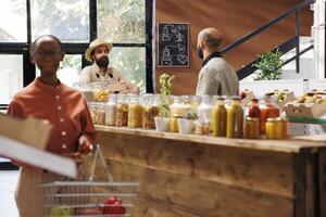 Black woman looks around in store while holding basket filled with food products. Focus on background are two men, one a farmer with boxes and another a store worker, interacting with each other. photo