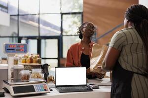 Blank white screen displayed on laptop while black woman with glasses receives her zero waste package. Isolated device with copy space for advertising or promotion of eco friendly bio food store. photo