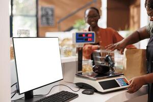 Seller using scale to measure organic products, with computer screen nearby having copy space display for bio food market commercial. Desktop computer at cashier desk showcasing blank mockup template. photo