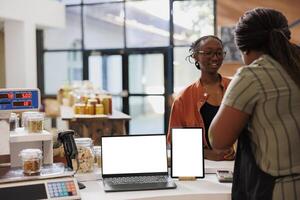 Customer at checkout counter with fresh, organic food products. Vendor promotes eco friendly, zero waste goods using laptop and tablet, white screen provides blank template for digital advertisement. photo
