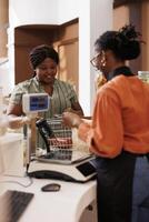African American woman buying eco friendly groceries from local store promoting sustainable lifestyle. Black female customer at checkout counter with shopping basket filled with fresh products. photo