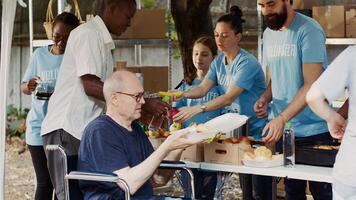 Senior old man in wheelchair receiving free food from group of multiethnic volunteers at hunger relief event. Friendly charity workers providing warm meals to the homeless and disabled people. photo