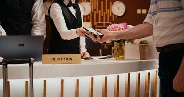 Senior man makes nfc payment with mobile phone at pos terminal, paying for room at five star hotel. Person arriving at reception desk and doing check in for vacation. Close up. Handheld shot. photo