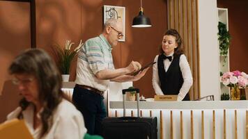 Elderly man signs digital forms for room check in at hotel front desk, talking to receptionist about leisure activities to ensure pleasant trip. Employee greeting visitor at reception. Handheld shot. photo