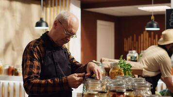 Portrait of smiling old zero waste vendor properly sealing bulk items decomposable glass containers. Elderly man in environmentally friendly local grocery shop arranging merchandise photo