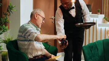 Elderly person paying for coffee cup via credit card in hotel lobby, ordering refreshment from bar and making pos transaction. Bellhop serving guest with beverage while he is waiting. photo