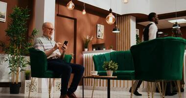 Retired traveller browses smartphone app in lounge area at five star holiday retreat, using online network to navigate on webpage. Senior client at hotel relaxing in lobby. photo