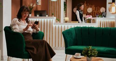 Old woman reads text messages after international flight, waiting for check in procedure at five star resort. Senior tourist browsing smartphone app in lounge area, relaxing in luxurious lobby. photo