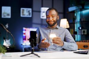 Online star doing technology vlog, showcasing dimmable smart light bulb to viewers. Friendly BIPOC content creator talking with subscribers. presenting IoT home automation photo