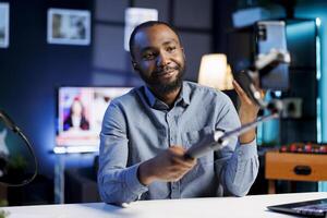 Cheerful entertainer using mobile phone attached to selfie stick to recording himself chatting with online audience. Online star discussing with fans during live broadcast stream from home studio photo
