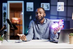 African american tech guru filming technology review of mini RGB lights device illuminating in different colors. Media star showing helpful production tool to subscribers photo