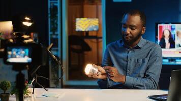 BIPOC content creator in dimly lit living room uses camera to film smart light bulb review for online streaming platforms. Media star hosts internet show, unboxing home automation product photo