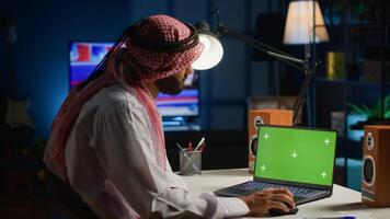 Arab man browsing on mockup laptop while working from home. Self employed freelancer checking emails on isolated screen device in apartment office with TV as background noise photo