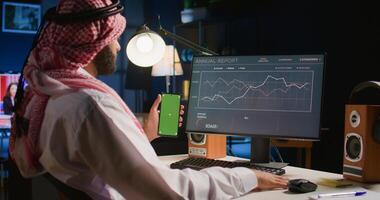 Man holding green screen smartphone while looking at business annual report graphs on computer. Self employed person with chroma key phone in hand performing tasks on device photo