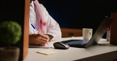 Man in apartment paying attention in elearning seminar, writing on notepad using pen. Middle Eastern homeschooling student listening to lesson while doing his homework tasks, close up photo
