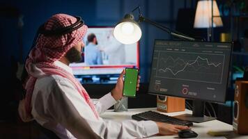 Remote worker holding green screen smartphone while looking at business annual report graphs on computer. Self employed freelancer with mockup phone in hand performing tasks on device photo