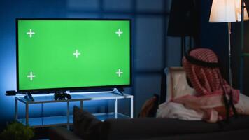 Arab man watching news broadcast on chroma key device while relaxing at home after hard day at work. Muslim person chilling on couch, looking at isolated screen TV channel photo