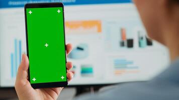 Business person holding greenscreen device at desk, looking at blank mockup display in coworking space. Woman showing mobile phone displaying isolated chromakey template, screen layout. Close up. photo