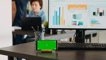 Empty workstation with greenscreen display on mobile phone app, desktop showing marketing analysis and reports in coworking space. Business office desk displaying smartphone screen with chromakey. photo