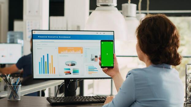 Employee looks at greenscreen on phone, working on business operations at desk. Person holding smartphone device with isolated display showing mockup template, blank layout screen. photo