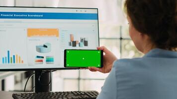Business specialist shows greenscreen on smartphone software display, looking at isolated chromakey template and working on computer at desk. Using blank mockup and finishing agency operations. photo