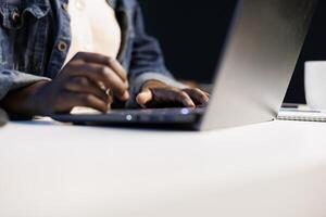 Close up of black person utilizing modern technology by typing on portable computer. Selective focus on hands of african american individual taking notes on laptop and surfing the net. photo