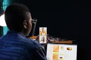 African American lady conferencing on smartphone while using personal computer to analyze charts. Black female freelancer grasping mobile device displaying ongoing call, online meeting. photo