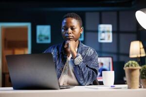 Female programmer sitting at home office desk and writing code on minicomputer, creating new software. African american woman working remotely, testing and looking for errors in program on laptop. photo