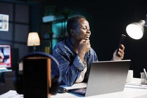 Excited black woman working remotely, using mobile device for calls. Cheerful female blogger uses a laptop and cellphone to browse the internet, check emails, and stay connected. photo