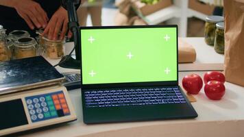 Vendor at register uses laptop running greenscreen display on device, young adult working at local organic supermarket looks at isolated copyspace. Seller waits for buyers at counter. photo