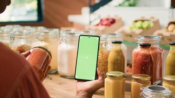 African american client uses greenscreen while she is looking at organic eco bulk products stored in reusable jars. Woman customer checks smartphone with isolated chromakey mockup display. photo