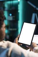 BIPOC man sitting on sofa, reading eBook from tablet in apartment using mockup tablet. Relaxed person enjoying interesting lecture on isolated screen device in neon lit living room photo