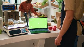 Shop owner works with greenscreen on laptop, sitting at register counter and waiting to serve customers in local zero waste supermarket. Vendor using blank display with chromakey mockup. photo