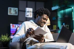 Entrepreneur working from living room, writing down company ideas while watching on laptop. Self employed man feeling ecstatic after coming up with startup proposal, using pen to note plan photo