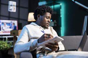 Entrepreneur working from RGB lit home, writing startup ideas on paper while watching on laptop. BIPOC man feeling delighted after coming up with entrepreneurship plan, using pen to note it down photo