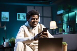 Smiling man at home taking break from work to write messages on phone. Joyous person holding cellphone talking with mates over online texting app before continuing project on laptop photo