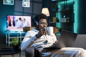 Happy man drinking coffee and writing emails at home office desk while listening music. Smiling remote worker doing b2b networking over internet, wearing headphones, enjoying hot beverage photo