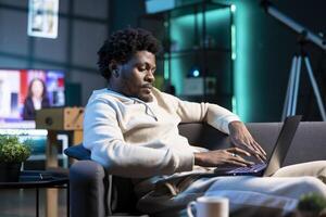 Relaxed remote worker checking his emails on laptop while laying on couch. BIPOC freelancer working from home in bright stylish cozy apartment personal office, typing on notebook photo