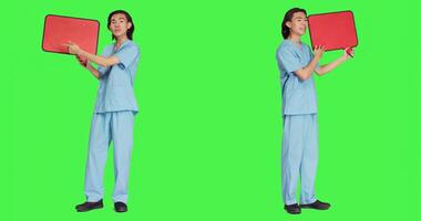 Young specialist holds speech bubble on camera with greenscreen template, holding billboard icon with blank backdrop. Healthcare employee showing small cardboard sign in studio. photo