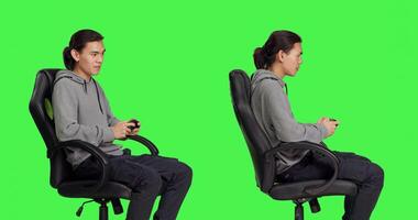 Young man playing videogames against greenscreen backdrop, sitting on chair and using controller on online gaming competition. Joyful relaxed guy having fun with rpg gameplay. photo
