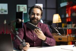 Cheerful online star doing technology reviews, excited to look at mini soundbar specifications. Influencer in studio delighted by high end music playing device, filming with smartphone on selfie stick photo