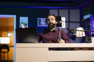 Happy influencer in home studio greeting audience, recording live broadcast using smartphone on selfie stick. Man filming q and a session with mobile phone, reading questions from notebook screen photo