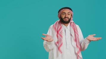 Muslim person being confused and unsure on camera, creating i dont know symbol and acting clueless over blue background. Young man in arab clothing being uncertain and shrugging. photo