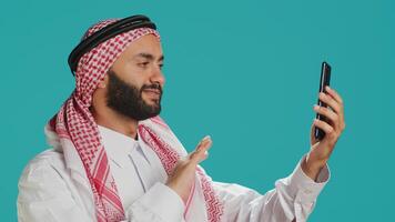 Arabic person engages in online conversation on his mobile device, wearing national costume. Young man with muslim headdress and thobe discussing on videoconference call in studio. photo
