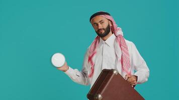 Arab guy sees wall clock to check time on camera, using timepiece to look at hour and minutes. Young person in traditional muslim clothing examining local timezone, standing over background. photo