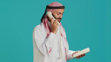 Arab guy talks on landline phone call in studio, answering call on retro telephone with cord for remote conversation. Young adult communicating with people on office phone line. photo